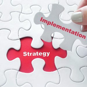 Close up of girl’s hand placing the last jigsaw puzzle piece wit Close up of girl's hand placing the last jigsaw puzzle piece with word Strategy Implementation as business concept