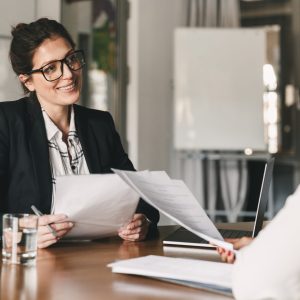 Portrait of smiling businesswoman holding resume and talking to Portrait of smiling businesswoman holding resume and talking to female candidate during corporate meeting or job interview - business, career and placement concept