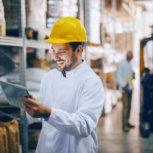 Young smiling Caucasian auditor in white uniform and with yellow Young smiling Caucasian auditor in white uniform and with yellow helmet on head using tablet for checking on goods in warehouse. In background worker standing next to forklift.